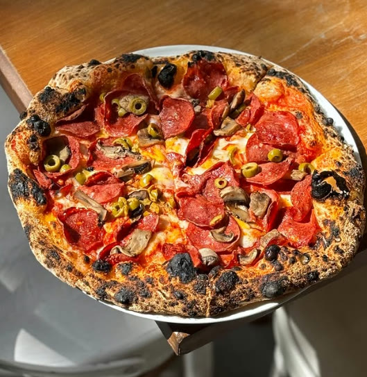Neapolitan pizza with pepperoni, mushrooms and olives — charred edges in sunlight